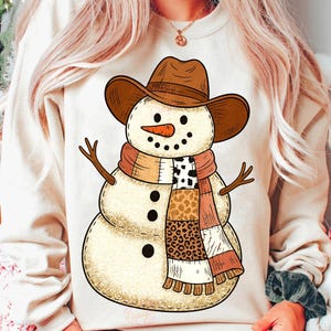 May include: A beige sweatshirt featuring a snowman design. The snowman wears a brown cowboy hat, a scarf with various patterns, and has a carrot nose and stick arms. Black dots form the eyes and mouth.