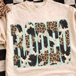 Pu&ograve; includere: T-shirt color crema con la scritta "RODEO" in un audace design con stampa leopardata bordata di azzurro. Sono visibili anche un berretto da baseball con stampa leopardata abbinato e scarpe da ginnastica a scacchi bianche e nere.