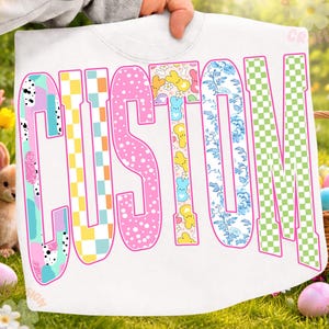 Può includere: Felpa bianca con la scritta "CUSTOM" a lettere grandi e colorate. Le lettere presentano vari motivi, tra cui pois, floreali e a scacchi. La felpa è tenuta da una persona, con uno sfondo sfocato di decorazioni pasquali.