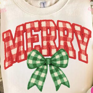 May include: Cream-colored sweatshirt with the word "MERRY" in red and white plaid letters. A green and white plaid bow is centered below the word. The sweatshirt is folded, and the design is centered on the front.