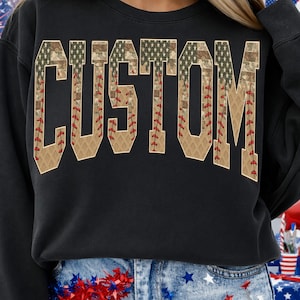 Puede incluir: Sudadera negra con la palabra "CUSTOM" en un diseño temático de béisbol. Las letras están rellenas con un patrón de camuflaje y un diseño de costura de béisbol. La sudadera se está usando.