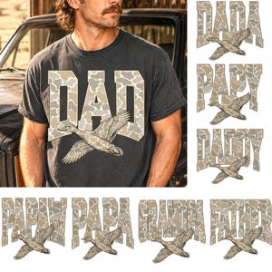 Op de afbeelding: Een antracietgrijs T-shirt met korte mouwen en het woord DAD in grote blokletters met een beige en bruin camouflagepatroon. Een illustratie van een vliegende wilde eend is over de tekst geplaatst. Inclusief PAPY, DADDY, PAPAW, PAPA, GRANDPA en FATHER.