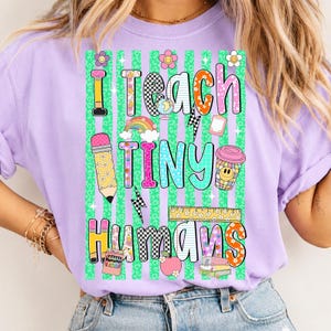 Puede incluir: Una camiseta de color morado claro con un estampado gr&aacute;fico que incluye el texto I TEACH TINY HUMANS. El dise&ntilde;o presenta un fondo de estampado de leopardo verde con rayas verticales, un l&aacute;piz, un globo terr&aacute;queo, un arco&iacute;ris, una taza, una regla y libros.