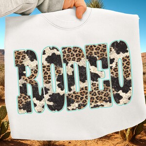 Pu&ograve; includere: Una felpa bianca con la scritta "RODEO" in lettere grandi e in grassetto. Le lettere hanno un contorno turchese e sono riempite con un mix di motivi leopardati e mucca. La felpa &egrave; tenuta contro un paesaggio desertico.