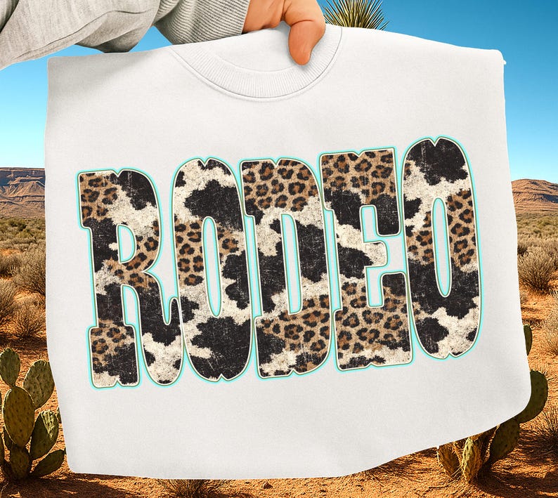 Pu&ograve; includere: Una felpa bianca con la scritta "RODEO" in lettere grandi e in grassetto. Le lettere hanno un contorno turchese e sono riempite con un mix di motivi leopardati e mucca. La felpa &egrave; tenuta contro un paesaggio desertico.