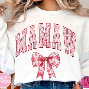 May include: White sweatshirt with the text "MAMAW" in red letters with a pink floral pattern. The letters are outlined in red and have a vintage collegiate style. A pink floral bow is below the text.