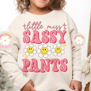 Op de afbeelding: Een licht beige sweatshirt met de tekst "little miss sassy pants" in roze letters met een retro lettertype. De tekst wordt omringd door drie gele smiley-gezichten met witte contouren in witte, madeliefje-achtige bloemen. De tekst "Daisy Design Crafts" staat in een cirkel met een witte madeliefje in het midden op de linker- en rechtermouw.