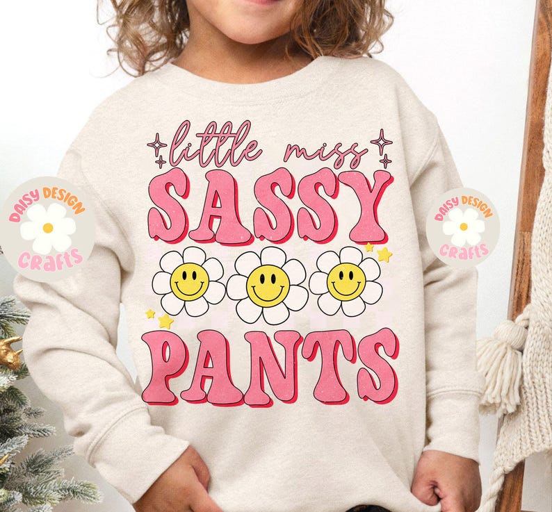 Op de afbeelding: Een licht beige sweatshirt met de tekst "little miss sassy pants" in roze letters met een retro lettertype. De tekst wordt omringd door drie gele smiley-gezichten met witte contouren in witte, madeliefje-achtige bloemen. De tekst "Daisy Design Crafts" staat in een cirkel met een witte madeliefje in het midden op de linker- en rechtermouw.