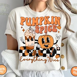 Peut inclure: T-shirt de couleur crème avec un motif rétro. Le design affiche les mots "PUMPKIN SPICE Everything Nice" en orange, ainsi qu'une tasse de café et une citrouille de dessin animé sur un fond à carreaux noir et blanc.