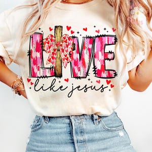 May include: Cream-colored t-shirt with the words "LIVE" in pink and red brushstrokes, a wooden cross, and a bow made of hearts. The phrase "like jesus" is written in cursive below, with red heart accents.