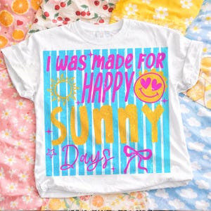 May include: White t-shirt with the text "I was made for Happy Sunny Days" in pink and yellow. The design includes a sun, smiley face, and bow on a blue striped background.