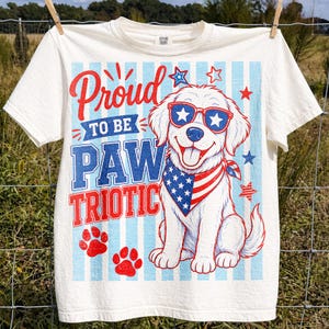May include: Off-white t-shirt with a patriotic design. The shirt features a cartoon dog wearing sunglasses and a bandana, with the text "Proud to be Paw Triotic" in red and blue. Red paw prints and stars complete the design.