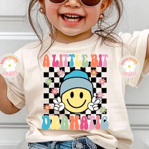 Op de afbeelding: Een wit T-shirt met een kleurrijk grafisch ontwerp met de tekst "A Little Bit Dramatic". De grafiek bevat een lachend gezicht met een blauwe hoed, een geruite achtergrond en bloemen.
