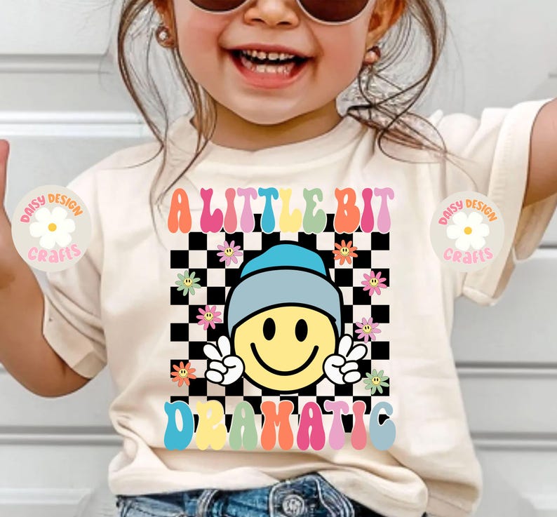 Op de afbeelding: Een wit T-shirt met een kleurrijk grafisch ontwerp met de tekst "A Little Bit Dramatic". De grafiek bevat een lachend gezicht met een blauwe hoed, een geruite achtergrond en bloemen.