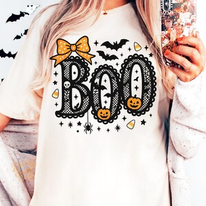 Puede incluir: Camiseta color crema con un dise&ntilde;o tem&aacute;tico de Halloween. La palabra "BOO" se muestra en una fuente decorativa, con un lazo, murci&eacute;lagos, calabazas y estrellas. El dise&ntilde;o es en negro y naranja.