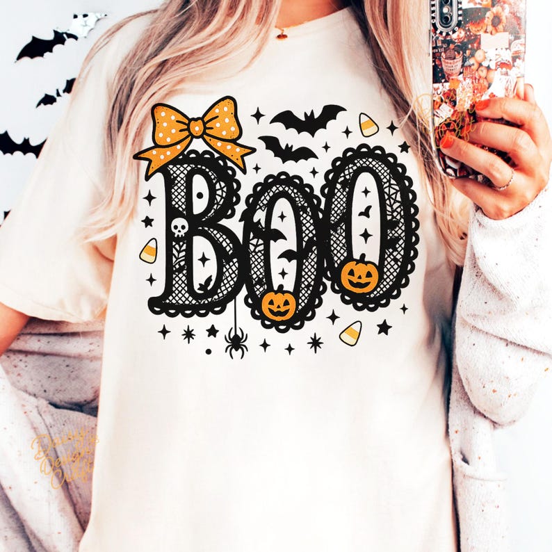 Puede incluir: Camiseta color crema con un dise&ntilde;o tem&aacute;tico de Halloween. La palabra "BOO" se muestra en una fuente decorativa, con un lazo, murci&eacute;lagos, calabazas y estrellas. El dise&ntilde;o es en negro y naranja.