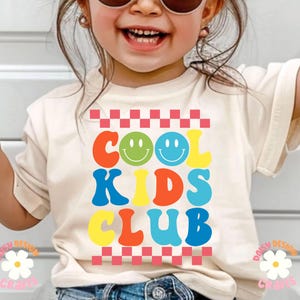 Op de afbeelding: Een wit T-shirt met een kleurrijke afbeelding waarop in een retro lettertype "Cool Kids Club" staat. De afbeelding bevat twee smiley-gezichten en een ruitpatroon.