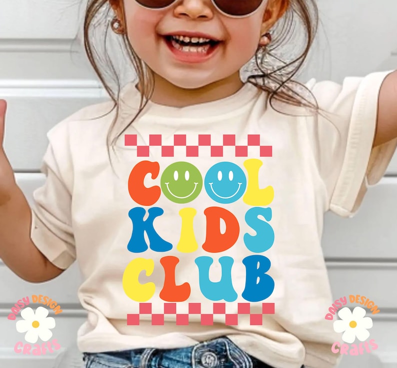 Op de afbeelding: Een wit T-shirt met een kleurrijke afbeelding waarop in een retro lettertype "Cool Kids Club" staat. De afbeelding bevat twee smiley-gezichten en een ruitpatroon.