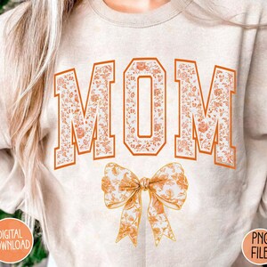 May include: A beige sweatshirt with the word "MOM" printed in orange and white floral letters. A large orange and white floral bow is below the word.