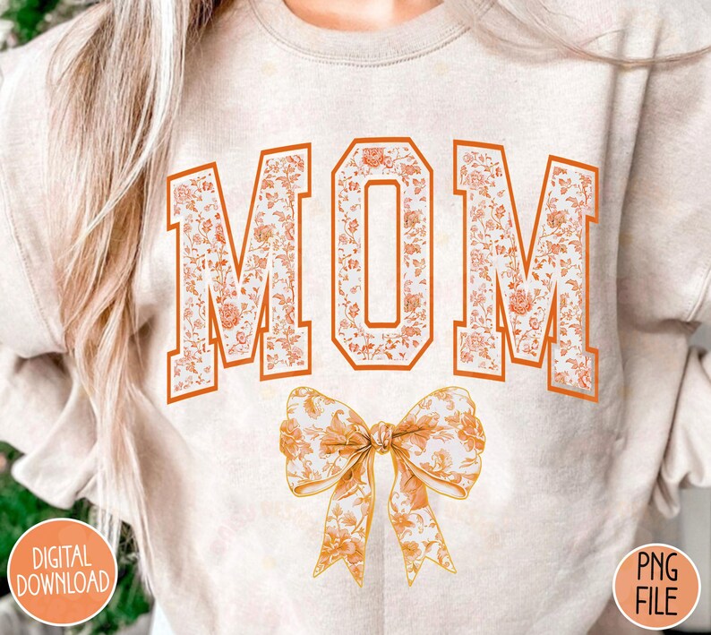 May include: A beige sweatshirt with the word "MOM" printed in orange and white floral letters. A large orange and white floral bow is below the word.