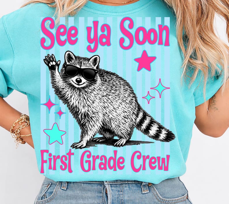 Op de afbeelding: Een lichtblauw T-shirt met de roze tekst "See ya Soon First Grade Crew". Het shirt heeft een wasbeer met een zonnebril die zwaait. Roze en turquoise sterren en lijnen versieren het shirt.