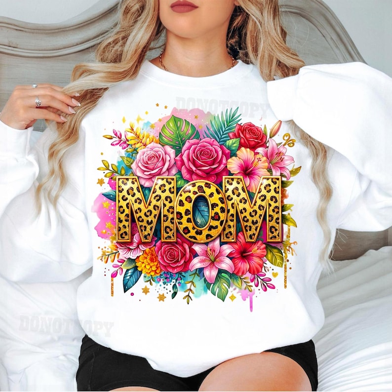 May include: White sweatshirt featuring the word "MOM" in gold leopard print, surrounded by a vibrant floral arrangement of pink roses, lilies, and tropical leaves. The design includes watercolor splashes and gold accents.