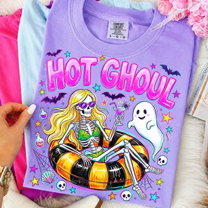 May include: A light purple t-shirt featuring a graphic of a skeleton with long blonde hair wearing sunglasses and a green bikini, lounging in a black and yellow striped pool float while holding a cocktail. The text reads HOT GHOUL.