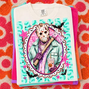 May include: A cream-colored t-shirt featuring a graphic of a hockey mask-wearing figure holding an axe and a pumpkin-themed drink. The design includes teal stripes, pink stars, and black bats within a scalloped oval frame. The shirt has a Comfort Colors label.