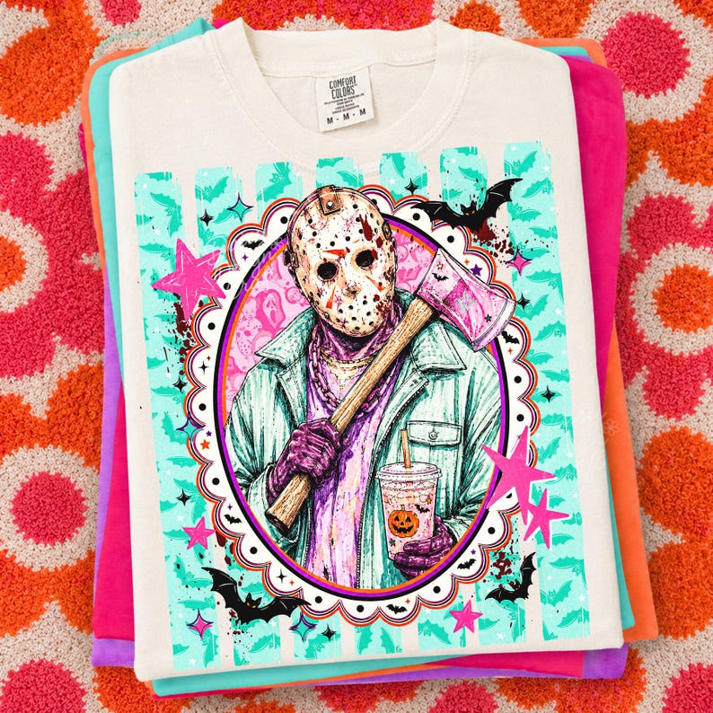May include: A cream-colored t-shirt featuring a graphic of a hockey mask-wearing figure holding an axe and a pumpkin-themed drink. The design includes teal stripes, pink stars, and black bats within a scalloped oval frame. The shirt has a Comfort Colors label.