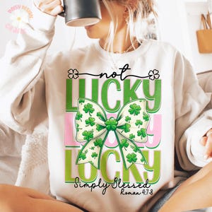 Może przedstawiać: Biała bluza z zieloną kokardką z koniczyny i tekstem "not lucky simply blessed Roman 4:7-8".