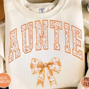 May include: A cream-colored sweatshirt with the word "AUNTIE" in orange letters with a floral pattern. A floral patterned bow is below the word.