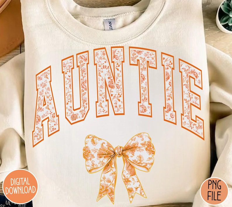 May include: A cream-colored sweatshirt with the word "AUNTIE" in orange letters with a floral pattern. A floral patterned bow is below the word.
