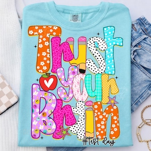 May include: Light blue Comfort Colors t-shirt with the words "Trust Your Brain" in colorful, playful lettering. The design includes an apple, pencils, stars, and smiley faces. The text "#Test Day" is at the bottom.
