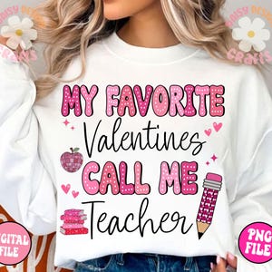 May include: White sweatshirt with pink polka dot lettering that reads "My Favourite Valentines Call Me Teacher". The text is surrounded by pink hearts, a pink apple, and a pink pencil.