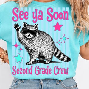Op de afbeelding: Een lichtblauw T-shirt met de tekst "See ya Soon Second Grade Crew". Het shirt heeft een wasbeer met een zonnebril die zwaait, met roze en turquoise sterren en strepen.