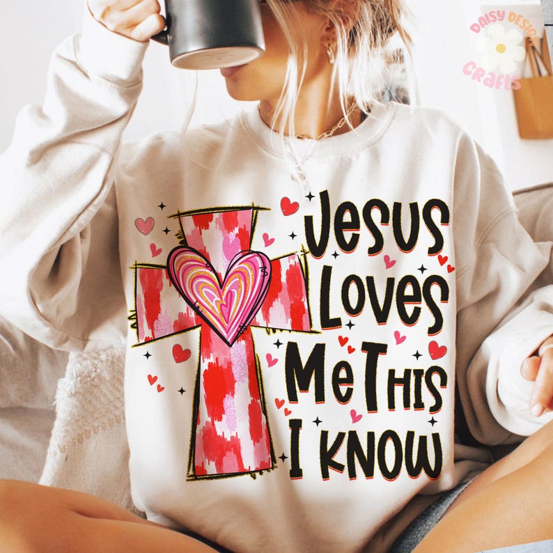 May include: White sweatshirt with a pink and red cross and the text "Jesus Loves Me This I Know" printed on it.