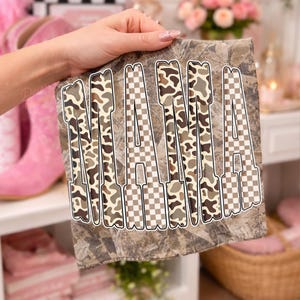 May include: A square, camouflage-patterned fabric with the word "MAMA" in large, bold letters. The letters alternate between a brown and tan camouflage design and a checkered pattern. The fabric is held up by a hand.