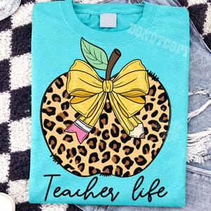 Puede incluir: Una camiseta de color turquesa con un gr&aacute;fico de una manzana con estampado de leopardo. La manzana est&aacute; adornada con un gran lazo amarillo y un l&aacute;piz. Debajo del gr&aacute;fico, el texto "Teacher life" est&aacute; impreso en una fuente cursiva negra.