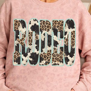 Pu&ograve; includere: Una felpa rosa chiaro con la scritta "RODEO" a lettere grandi e in grassetto sul davanti. Le lettere hanno un contorno turchese e sono riempite con un mix di motivi leopardati e mucca. La felpa ha uno scollo tondo.