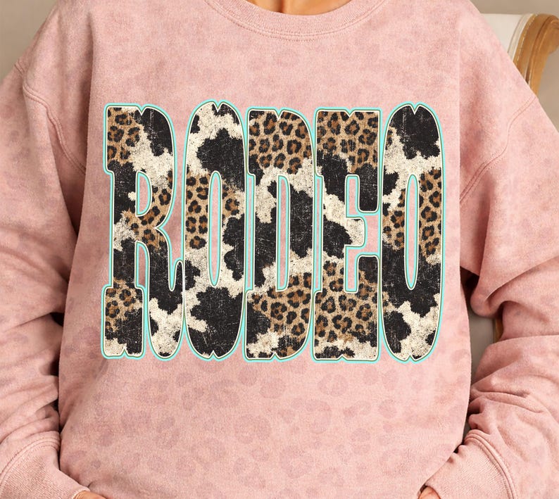 Pu&ograve; includere: Una felpa rosa chiaro con la scritta "RODEO" a lettere grandi e in grassetto sul davanti. Le lettere hanno un contorno turchese e sono riempite con un mix di motivi leopardati e mucca. La felpa ha uno scollo tondo.