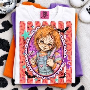 May include: A white t-shirt featuring a graphic of a doll with orange hair, a stitched face, and a knife, framed by a scalloped oval. The design includes pink and orange vertical stripes, bats, stars, and a lollipop. The neck label reads Comfort Colors.