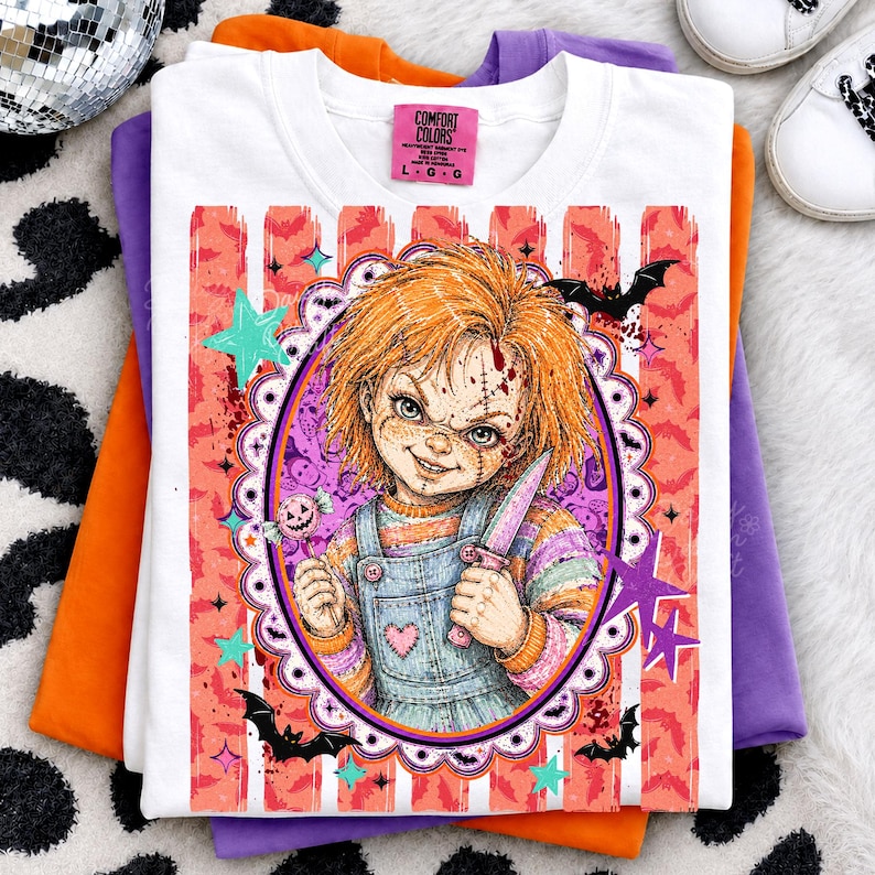 May include: A white t-shirt featuring a graphic of a doll with orange hair, a stitched face, and a knife, framed by a scalloped oval. The design includes pink and orange vertical stripes, bats, stars, and a lollipop. The neck label reads Comfort Colors.