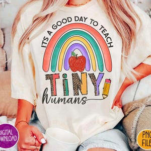 K&ouml;nnte beinhalten: Ein wei&szlig;es T-Shirt mit einem Regenbogen-Design und dem Text "Es ist ein guter Tag, um kleine Menschen zu unterrichten". Der Regenbogen hat einen roten Apfel in der Mitte.