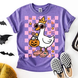 Op de afbeelding: Paarse t-shirt met een cartoon gans met een zonnebril en een kroon, met een pompoen. De gans staat op een roze en paarse geruite achtergrond met Halloween-afbeeldingen. Geruite Vans-schoenen staan onderaan.