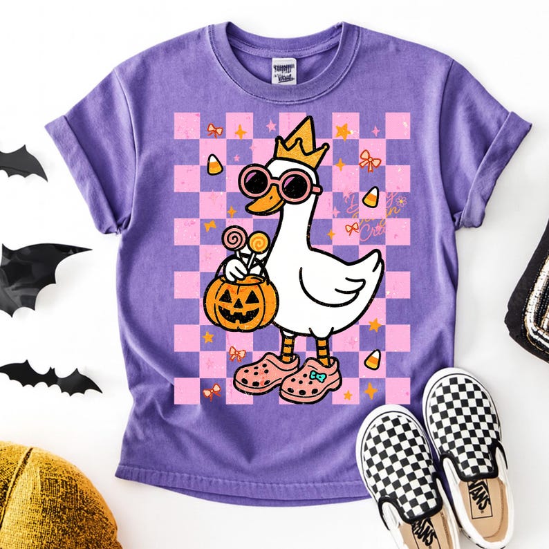 Op de afbeelding: Paarse t-shirt met een cartoon gans met een zonnebril en een kroon, met een pompoen. De gans staat op een roze en paarse geruite achtergrond met Halloween-afbeeldingen. Geruite Vans-schoenen staan onderaan.