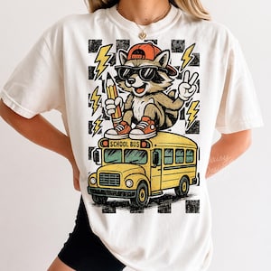 Puede incluir: Una camiseta blanca con un gr&aacute;fico de un mapache con gafas de sol y una gorra naranja, sosteniendo un l&aacute;piz y haciendo el signo de la paz. El mapache est&aacute; sentado sobre un autob&uacute;s escolar amarillo, sobre un fondo de cuadros negros y blancos.
