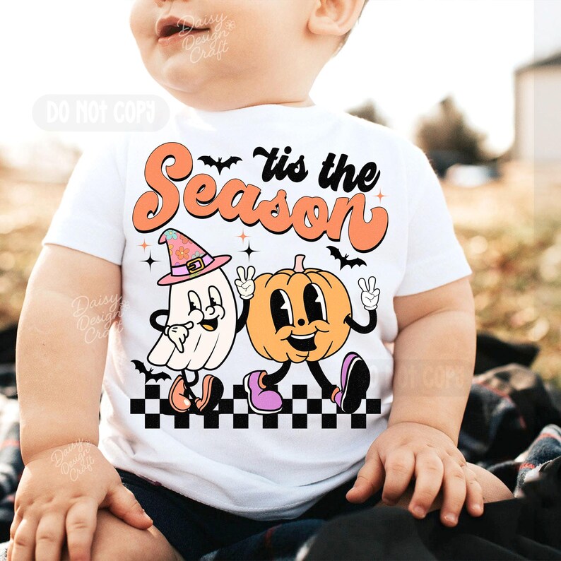May include: White t-shirt with a retro Halloween design. The shirt features the text "'tis the Season" above a cartoon ghost wearing a witch hat and a pumpkin, both giving peace signs. Black bats and a checkered pattern complete the design.