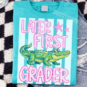 以下が含まれることがあります： 漫画のアリゲーターのグラフィックとピンク色の「LATER FIRST GRADER」という言葉がプリントされた、ティール色のTシャツ。デザインにはピンクの星と水色のストライプが含まれています。このシャツは柔らかい素材で作られています。