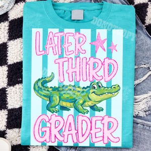 以下が含まれることがあります： ピンクのグリッター文字で「LATER THIRD GRADER」と書かれた、ターコイズ色のTシャツ。テキストの下には漫画のアリゲーターが描かれ、上にはピンクの星が描かれています。シャツは水色のストライプの背景です。
