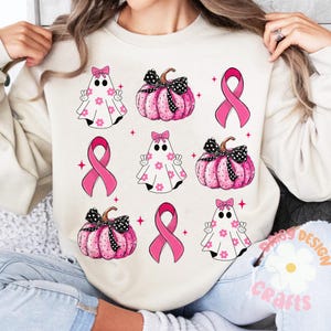 May include: A white sweatshirt with a pink ribbon, ghosts, and pumpkins. The design features a pink ribbon, a ghost with a pink bow, and a pumpkin with a pink bow. The design is repeated throughout the sweatshirt.